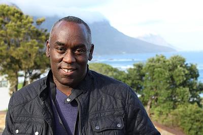 Alex Wheatle smiling at the camera with trees, mountains and the sea in the background